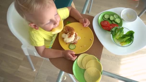 Menino safado brincando com hambúrguer ingredientes queijo sentado à mesa — Vídeo de Stock