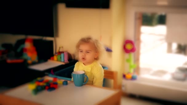 Маленькая кудрявая девочка пьет из голубой чашки в детской комнате. 4K — стоковое видео