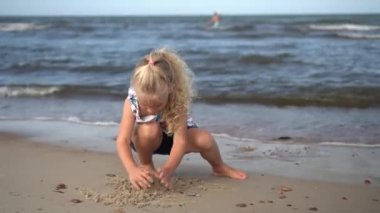 Sarışın kız sahilde kumla oynuyor. Deniz suyu dalgaları ve bulanık insanlar