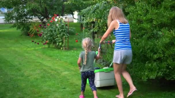 Мама учит дочку стричь траву газонокосилкой в летнем саду — стоковое видео