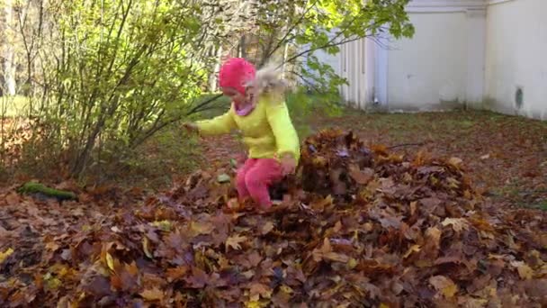 Lächelndes blondes Mädchen springt auf bunten Laubhaufen im Hinterhof des Hauses — Stockvideo