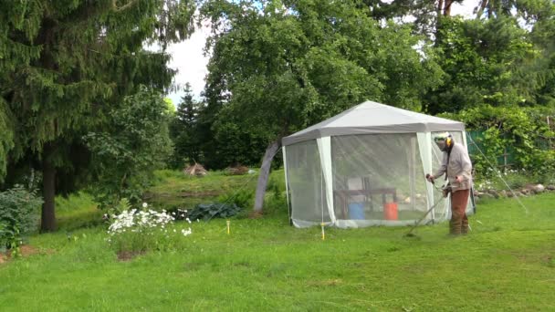 Κηπουρός άνθρωπος τελειώματα χορτοτάπητα με το θεριστή κοντά καλοκαίρι κληματαριά bower — Αρχείο Βίντεο