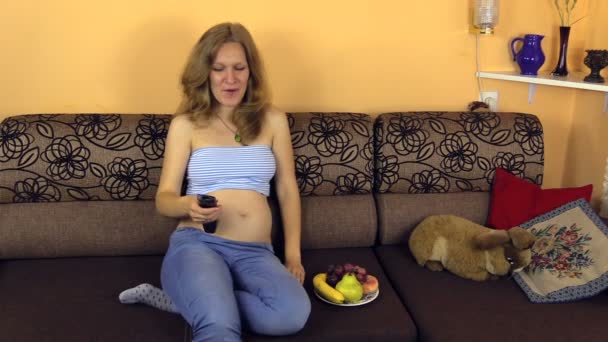 Heureuse femme enceinte caresse son ventre manger du raisin et regarder la télévision — Video