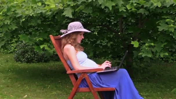 Pregnant woman with hat work with laptop sitting on wooden chair — Stock Video