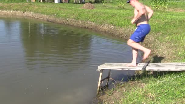 Людина в шортах і капелюсі біжить по дерев'яному мосту і стрибає в озерній воді — стокове відео