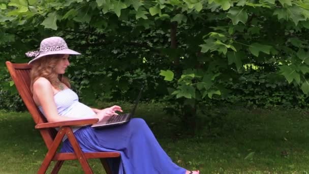 妊娠中の女性の帽子とラップトップ座る椅子緑豊かな公園に上で動作します。 — ストック動画