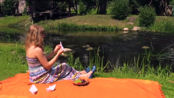 Chica hacer barco de papel sentarse en cuadros cerca de riachuelo ondulado en verano — Vídeos de Stock
