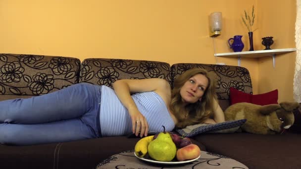Žena leží na gauči, palm břicho, stiskněte tlačítko dálkového ovládání tv. — Stock video