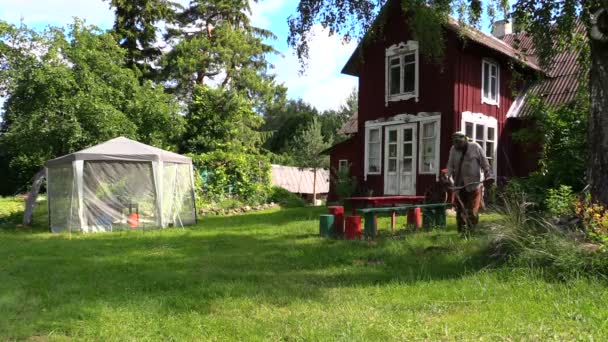 Gärtner mähte mit Trimmer Gras in der Nähe von Bauernhaus und Laube — Stockvideo