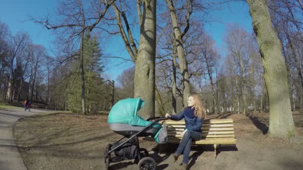 年轻的母亲抱着婴儿车坐在公园的长凳上。4 k — 图库视频影像