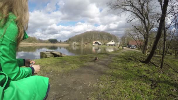 Blonde vrouw met groene vacht feed eend vogels in de buurt van lake. 4k — Stockvideo