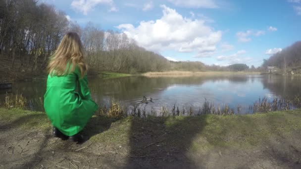 Mulher loira com casaco verde alimentando pássaros de pato perto do lago. 4K — Vídeo de Stock