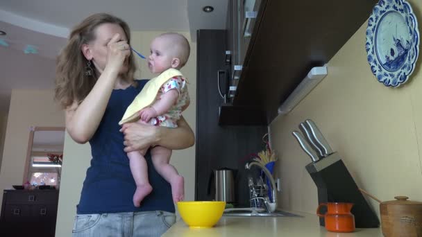 Жінка годує дочку дитиною кашею, тримаючись за руку на кухні — стокове відео