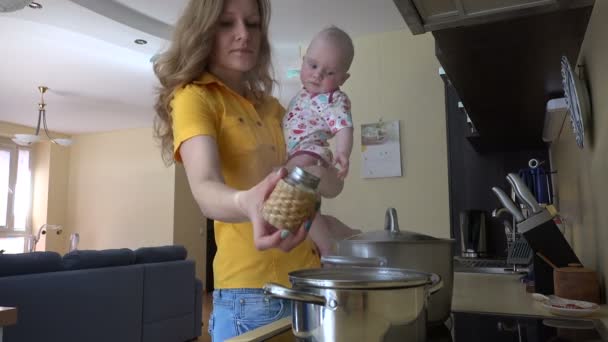 Mujer joven sostiene bebé niña, preparar la comida en la cocina. 4K — Vídeos de Stock