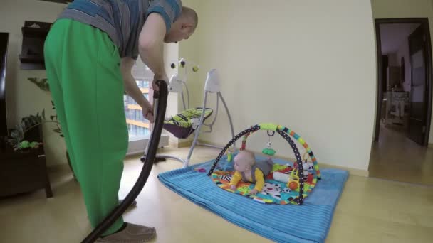 Man reiniging van de vloer met hoover en nieuwsgierig baby op spelen mat. 4k — Stockvideo