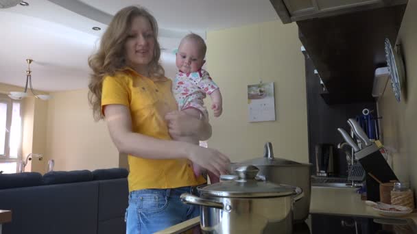 Молодая мама держит малышку, готовит еду на кухне. 4K — стоковое видео