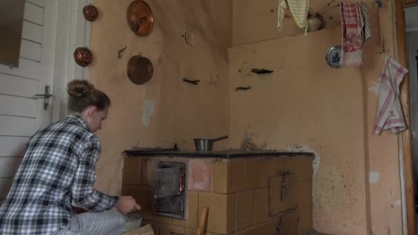 Young villager woman put some firewood in rural kitchen stove. 4K — Stockvideo