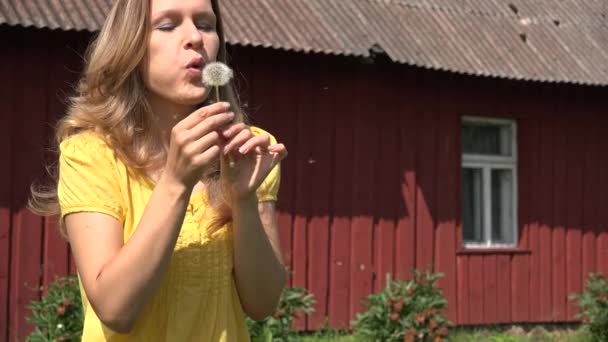 Mulher soprar dente de leão perto da casa rural e sementes voando no ar. 4K — Vídeo de Stock