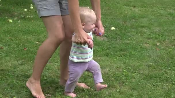 Lindo bebé dando los primeros pasos en el césped verde, madre cogida de la mano apoyando enseñando a caminar . — Vídeo de stock