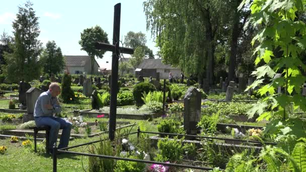 Jeune homme assis wince sur le banc près de la tombe de la femme au cimetière de campagne. 4K — Video