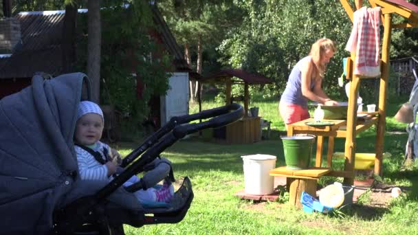 Bebek bebek arabası kraker yemek ve köy bahçesinde çamaşır yıkama anne kadın bakmak. 4k — Stok video