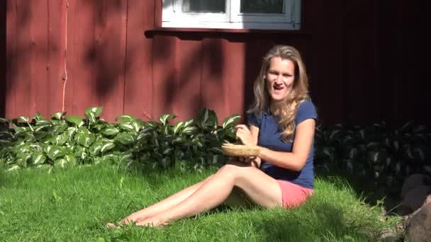 Σέξι γυναίκα με σορτς κάθεται κοντά σε αγροτικό ξύλινο σπίτι τρώγοντας βατόμουρο μαύρο βατόμουρο και χαμόγελο. 4K — Αρχείο Βίντεο