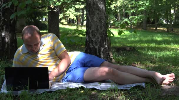 Un hombre serio trabajando con un ordenador portátil tirado bajo los árboles en el parque. 4K — Vídeos de Stock