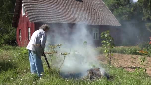 A mulher agricultora aposentada queima grama e se recusa no fogo no prado no quintal da casa da aldeia rural. 4K — Vídeo de Stock