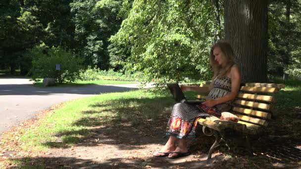 Элегантная женщина в платье сидит на скамейке и работает над ноутбуком в парке. 4K — стоковое видео