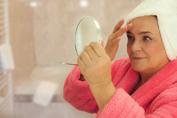 Portrait of an attractive middle aged woman looking into a mirror in the bathroom — Stock Photo, Image