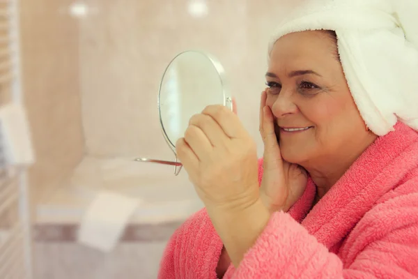 Portrait of an attractive middle aged woman looking into a mirror in the bathroom — Stock Photo, Image