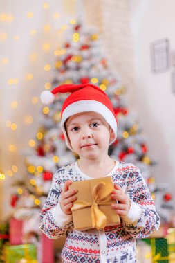 Noel ağacında hediyeli bir çocuk. - Evet. Çocuk ve Noel ağacı. Sevimli bebek. Tatil günü. Yeni yıl ve Noel hakkında bir makale. Renkli ladin. Çelenk. Tatil dekorasyonu ve dekorasyonu. Noel Baba şapkası. Yeni yıl hediyelerini seçmekle ilgili makale