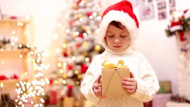 Niño en el árbol de Navidad con un regalo. .. El chico sacude el regalo. Lindo bebé. Vacaciones. Artículo sobre año nuevo y Navidad. Picea colorida. Decoración y decoración navideña. Sombrero de Santa. Artículo sobre — Vídeo de stock