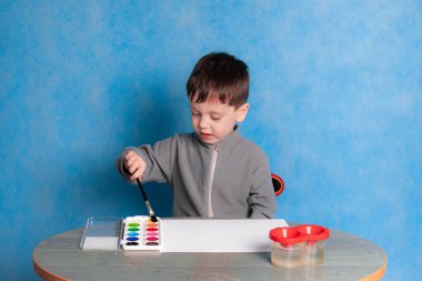 Çocuk tek renkli bir arkaplan çiziyor. Bir çocukla resim reklamı yapmak. Sade arka planda boyaları olan bir çocuk. Mutlu bir çocukluk. Hobi çizimi. Çocuk çizimi. Çocuklar için resimler. Evde, karantinada dersler. Çocukların boş vakitleri. Katı mavi