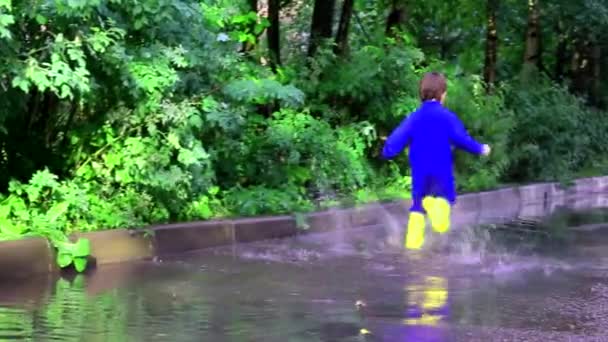 De jongen springt in een plas. Een jongen met rubberen laarzen. Gelukkige jeugd. Zomer. Zomerpoelen. Vers na de regen. Kinderplezier. — Stockvideo