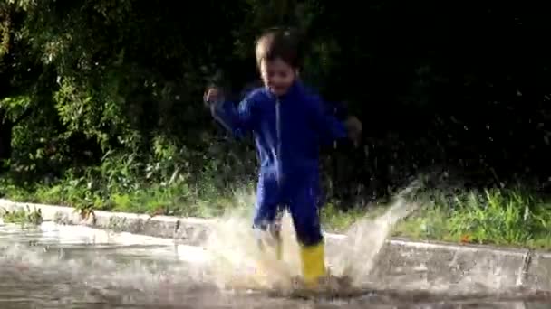 その少年は水たまりに飛び込む。ゴムのブーツを着た少年。幸せな子供時代。夏だ。夏の水たまり。雨の後に新鮮な。子供の楽しみ. — ストック動画