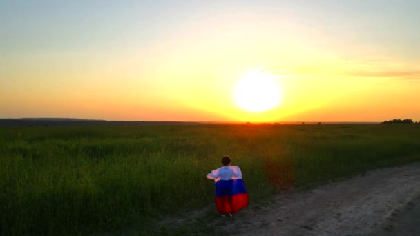 Ένα παιδί σε ένα χωράφι με ρωσική σημαία το ηλιοβασίλεμα. Βραδινό φως. Καλοκαιρινό τοπίο στο ηλιοβασίλεμα. Πατριωτισμός. Η σημαία της Ρωσίας κυματίζει στον άνεμο. — Αρχείο Βίντεο