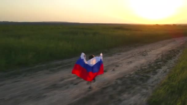 Ein Kind auf einem Feld mit einer russischen Flagge bei Sonnenuntergang. Abendlicht. Sommerlandschaft Feld bei Sonnenuntergang. Patriotismus. Die Flagge Russlands weht im Wind. — Stockvideo