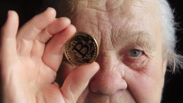 An elderly woman, a grandmother, is holding a bitcoin coin in her hands and leans it to her eye — Stock Video