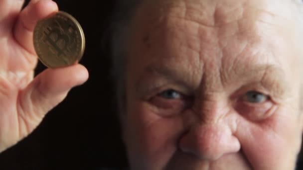 An elderly woman, a grandmother, is holding a bitcoin coin in her hands and leans it to her eye — Stock Video