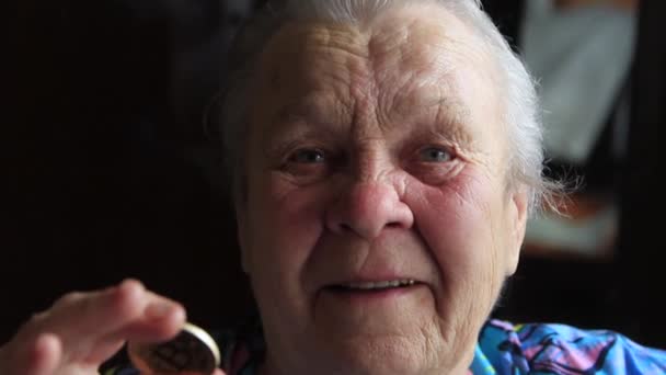 An elderly woman, a grandmother, is holding a bitcoin coin in her hands and leans it to her eye — Stock Video