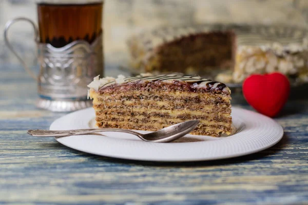 Un morceau appétissant de gâteau avec des noix, de la crème et du chocolat, du thé dans un verre et un cœur rouge sur un fond en bois bleu clair. Concept de célébration de la Saint-Valentin. — Photo