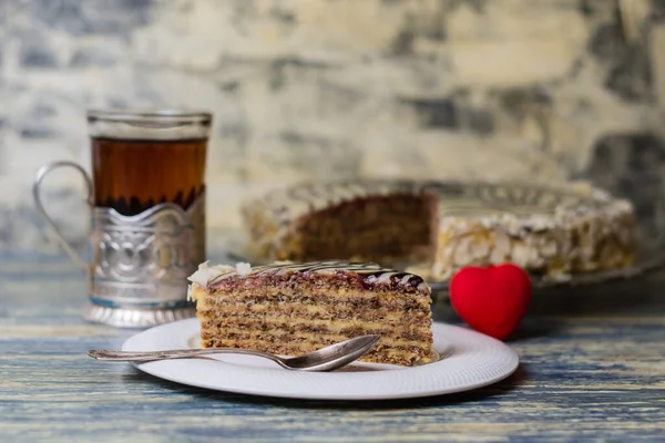 Un morceau appétissant de gâteau avec des noix, de la crème et du chocolat, du thé dans un verre et un cœur rouge sur un fond en bois bleu clair. Concept de célébration de la Saint-Valentin. — Photo