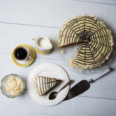 Çikolatalı, kremalı, bademli gurme pastası, açık ahşap arka planda bir fincan espresso ve süt, kare format..