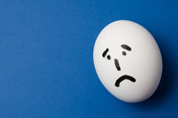 Egg with a sad face, on blue background with copy space.