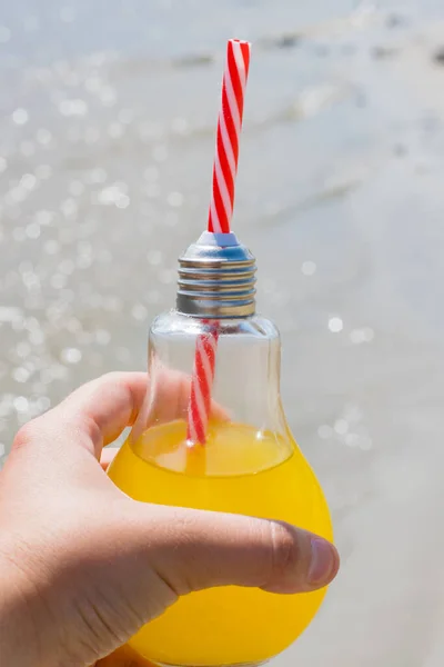 Świeże lub koktajl w szklanej butelce ze słomką zbliżenie w żeńskiej dłoni na tle plaży i wody, pionowy format. Koncepcja podróży i rekreacji. — Zdjęcie stockowe