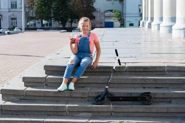 在城市的台阶上 十几岁的女孩拿着柠檬水和一个塑料杯子 还有一根稻草和一辆摩托车 — 图库照片