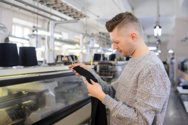 Endüstriyel örgü makinelerin yanında siyah iplikli kumaş parçalarını analiz eden genç bir adam. 