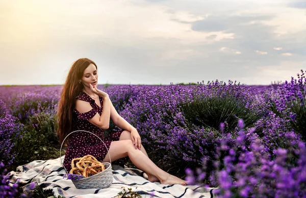 Portrait of a young woman in violet dress with dots having a picnic with a basket full of cookies and bagels in a lavender field. Lavender flowers bloom