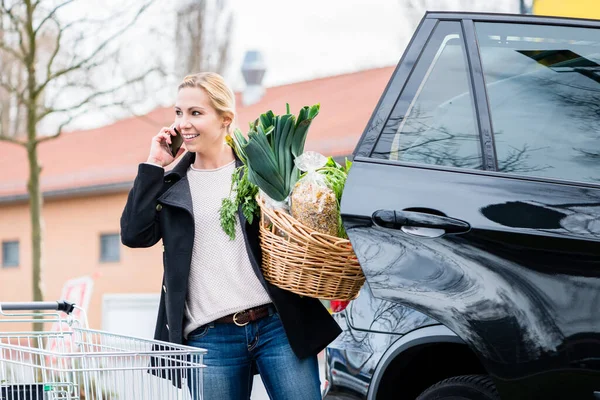 Wanita menggunakan telepon sambil menyimpan bahan makanan di bagasi mobil — Stok Foto